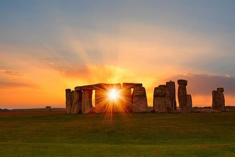 The sun sets behind Stonehenge, casting a golden glow through the iconic stone circle, highlighting its ancient mystery and beauty.