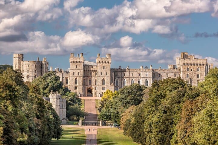 Windsor Castle seen from the Long Walk, framed by trees, highlighting the majestic structure standing proudly against a backdrop of clouds.