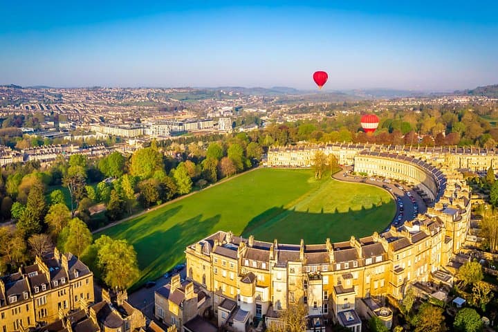 A breathtaking aerial view of Bath’s iconic Royal Crescent, with hot air balloons floating over the city on a clear day.