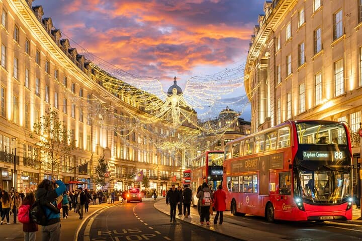 Regent Street illuminated with festive lights and bustling with red double-decker buses at sunset, capturing the lively atmosphere of central London.
